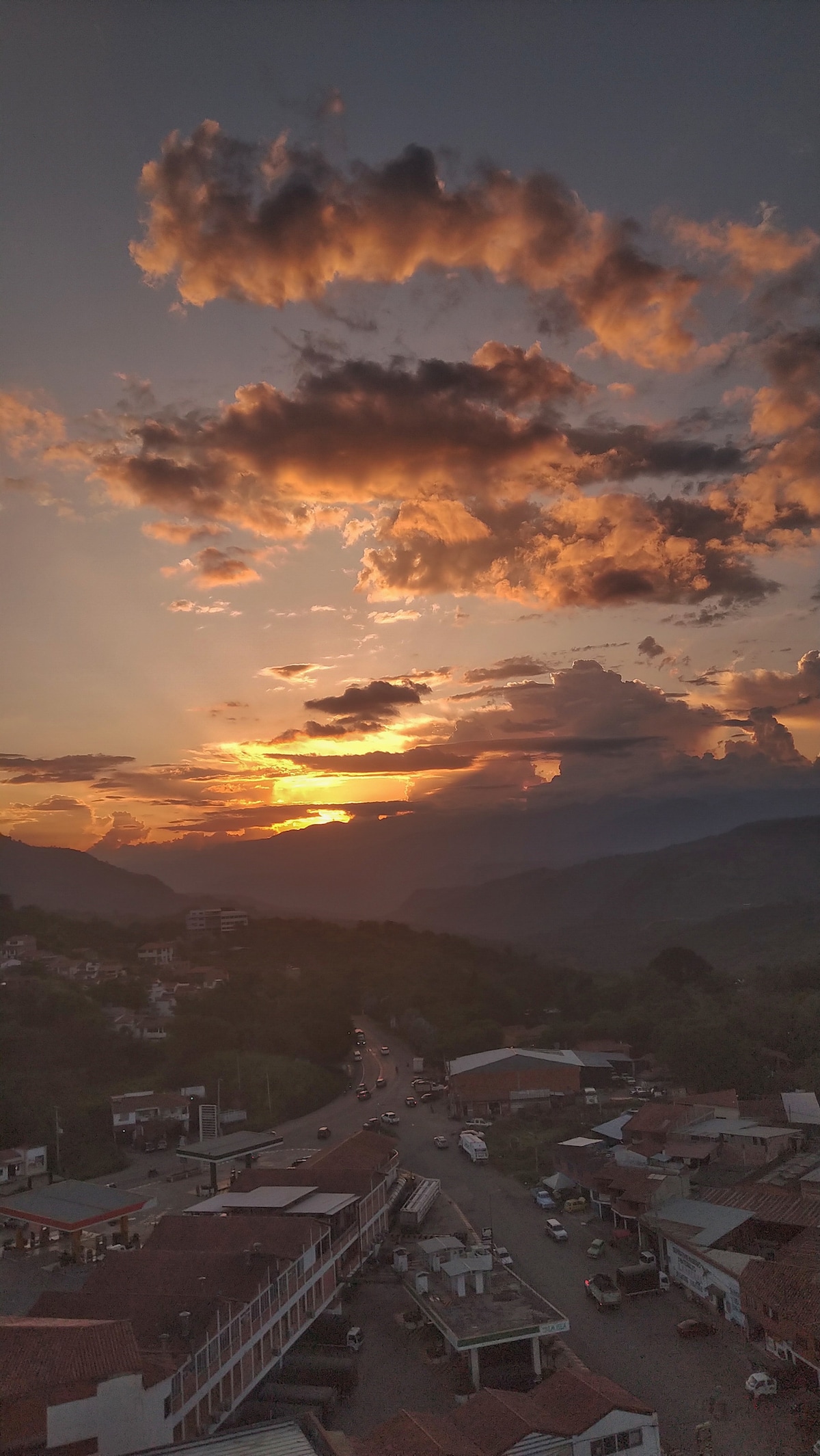 A vibrant sunset paints the sky in warm hues, illuminating the rolling hills and distant mountains. The view captures a winding road that meanders through the landscape, with rooftops of nearby buildings visible in the foreground.