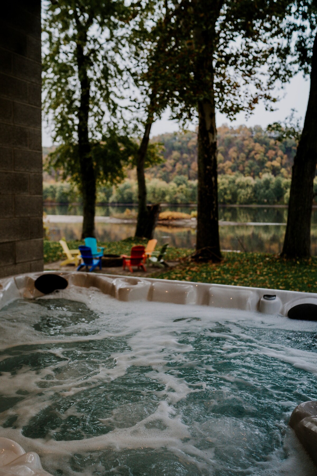 A hot tub with bubbling water is situated beside a river, offering scenic views of trees and rolling hills in the background. Colorful chairs in red and blue are positioned nearby, enhancing the outdoor relaxation space.