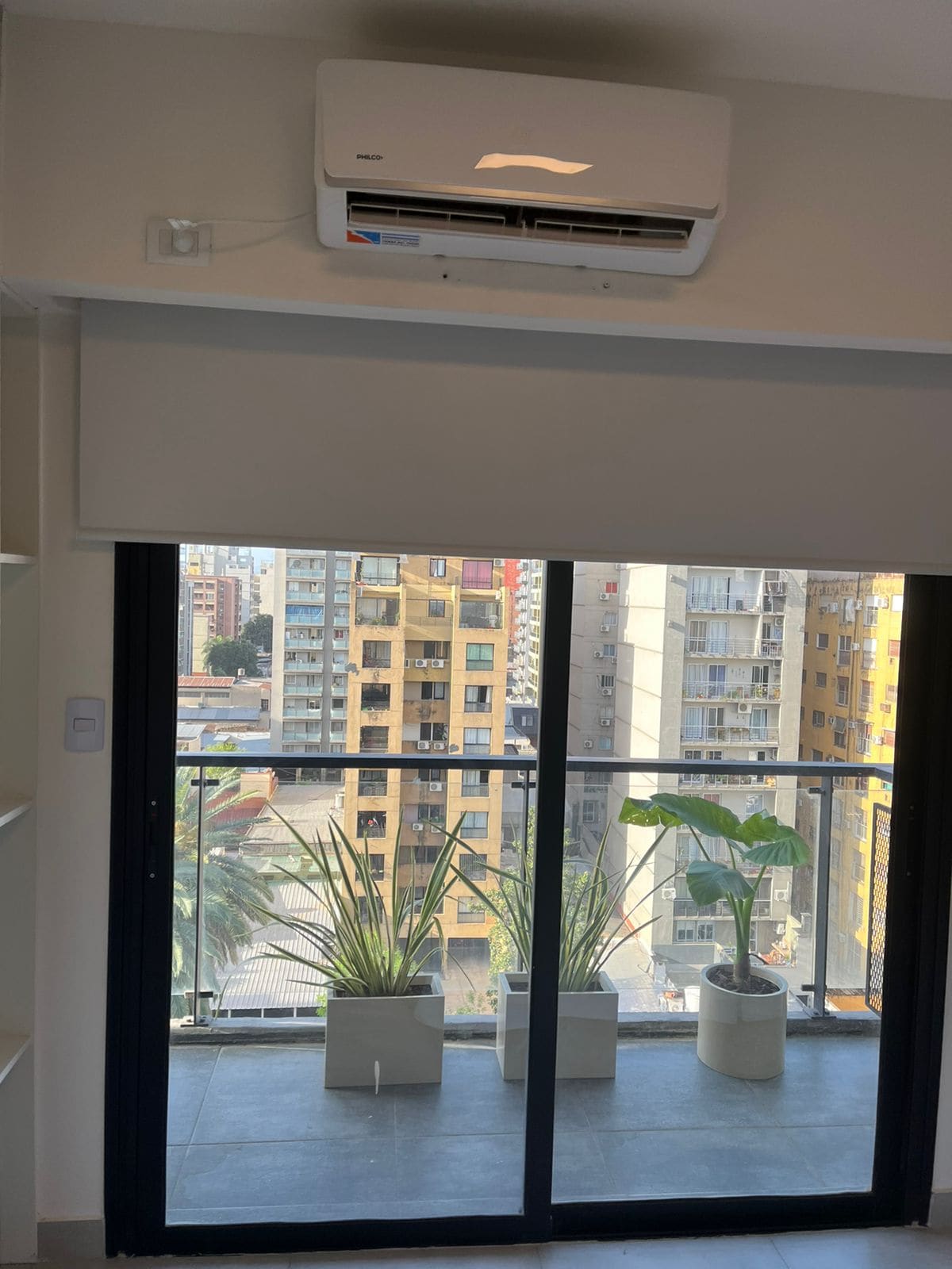 A bright space is featured with a sliding glass door opening to a small balcony. Potted plants are positioned on the balcony floor, adding a natural touch. An air conditioning unit is mounted above the door, providing climate control for the room.