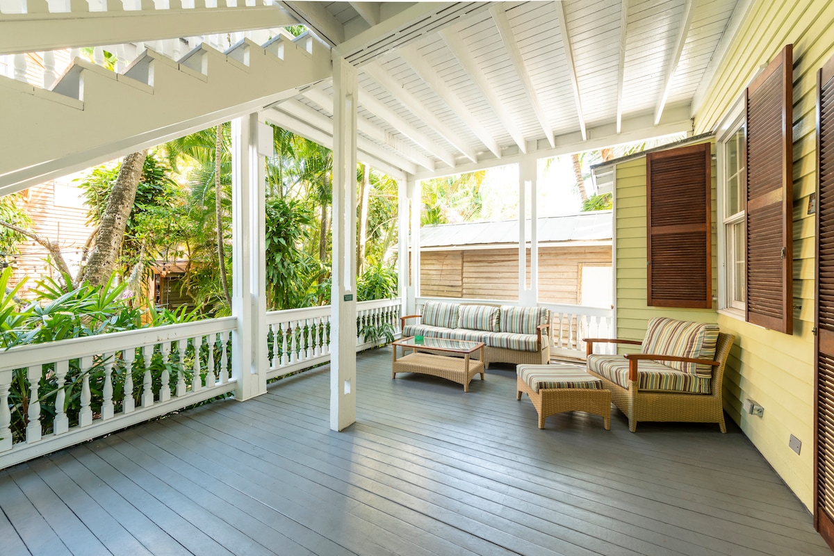 A spacious outdoor porch is decorated with comfortable furniture, including a couch with striped cushions and a wooden coffee table. Lush greenery surrounds the area, providing a natural backdrop, while light filters through the overhead beams and the charming lattice work along the railing.
