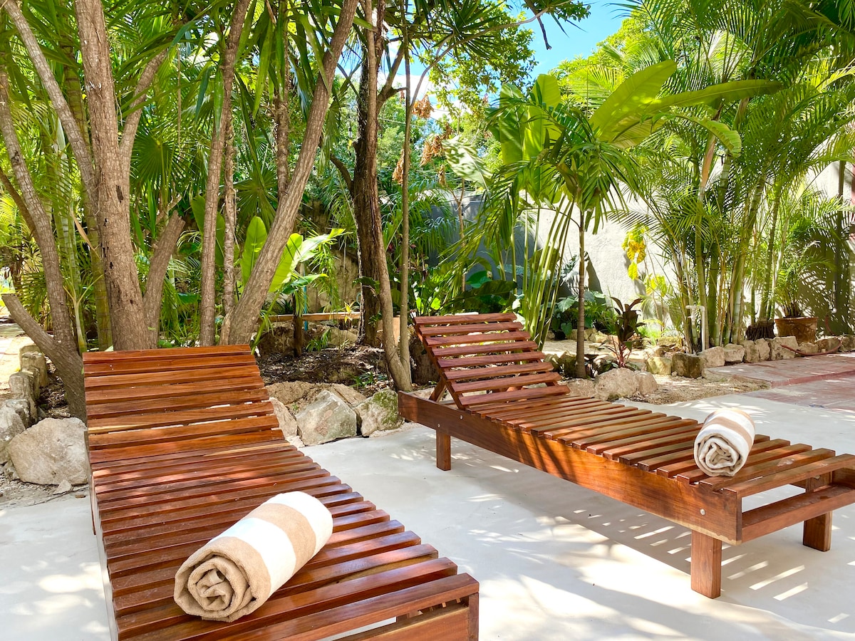 Two wooden lounge chairs are positioned under lush tropical trees, creating a serene setting. Each chair is adorned with a rolled towel, inviting relaxation in the surrounding greenery and soft sunlight.