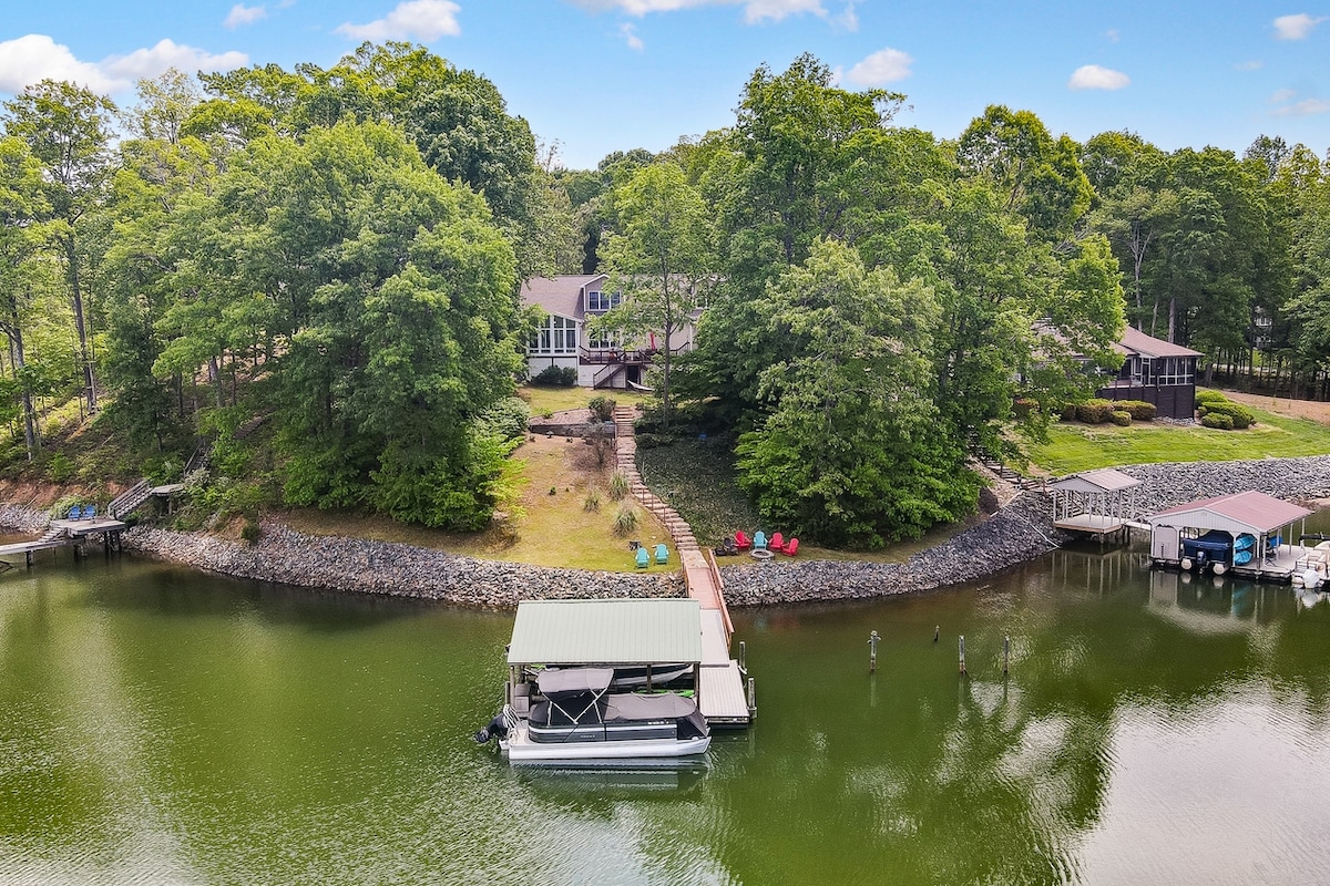 An aerial view showcases the serene lakeside property surrounded by lush greenery. The calm water reflects the vibrant trees, while a spacious lawn and pathways lead to the large boat dock and seating areas, creating an inviting outdoor space for relaxation.
