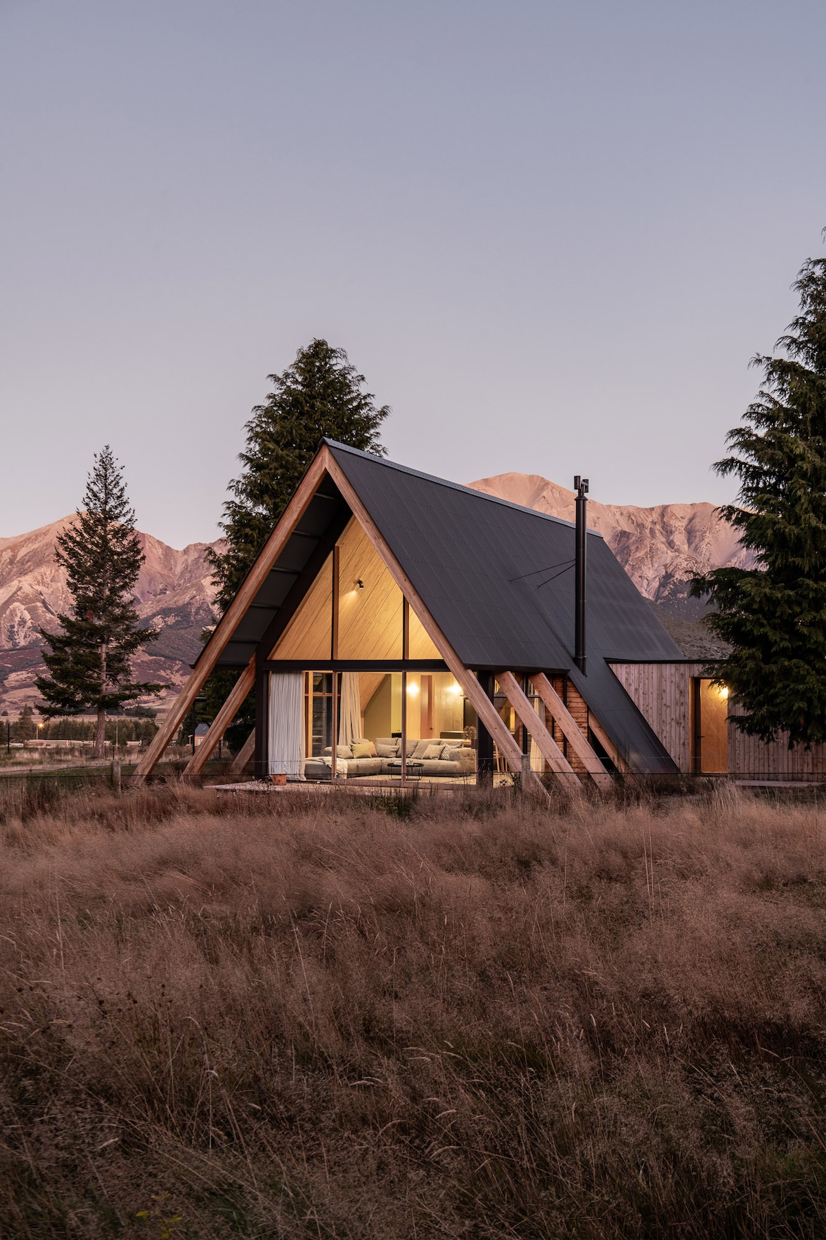 The A-frame house is set against a mountainous backdrop, with large glass windows revealing a warm interior. The exterior features wooden accents and a sloping roof, surrounded by tall grass and trees, creating a natural integration with the alpine environment.