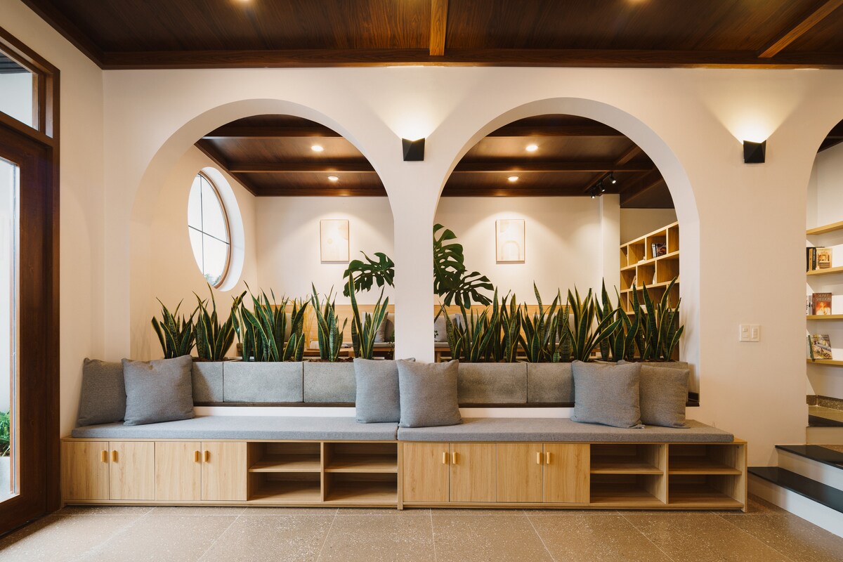 A modern seating area features a built-in bench adorned with soft grey cushions. Tall plants are arranged within alcoves, and archways provide an open, inviting passage. Natural light filters through round windows, enhancing the warm wooden details of the ceiling.