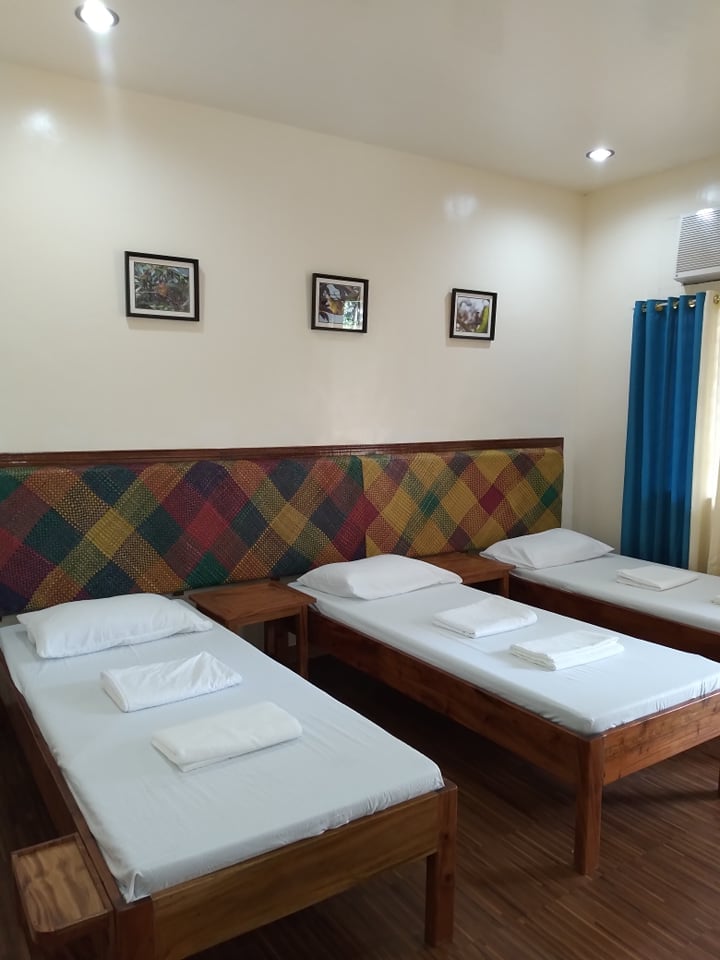 The room features three wooden beds with white linens and neatly folded towels. A colorful patterned headboard adds visual interest. Natural light enters through a window dressed with blue curtains, and framed artwork is displayed on the walls.