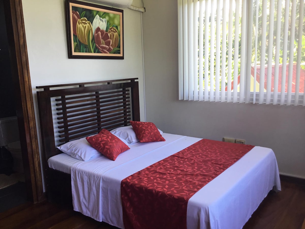 A bright sleeping area features a spacious bed dressed with a white base and a red decorative runner. Two red accent pillows are positioned at the head of the bed. Vertical blinds allow natural light to flow in from a nearby window, highlighting a floral artwork on the wall.