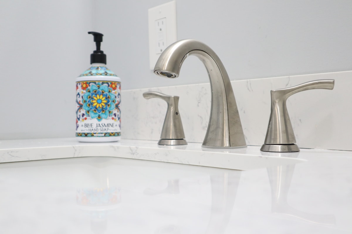 A modern bathroom sink area features sleek metal faucets, complemented by a decorative hand soap dispenser adorned with vibrant floral patterns. The countertop showcases a smooth white surface with subtle marbling, providing a clean and fresh appearance.