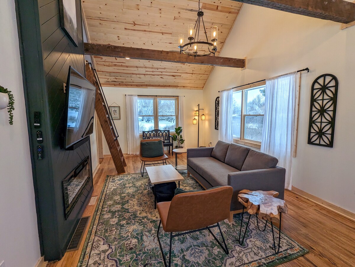 The living room showcases a high vaulted ceiling with original hand-hewn beams and a modern chandelier. A spacious sofa is positioned across from a sleek fireplace, complemented by a unique side table and decorative area rug. Large windows allow natural light to fill the space.