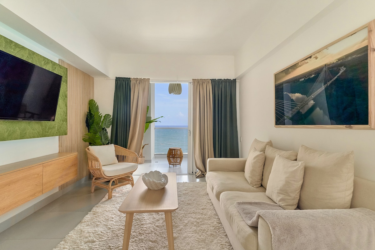 A sunlit living area features a large, cozy sofa facing a mounted television. Natural light spills in through large sliding doors that open to a sea view. A small wooden coffee table is positioned on a plush rug, alongside a comfortable chair and decorative plant.
