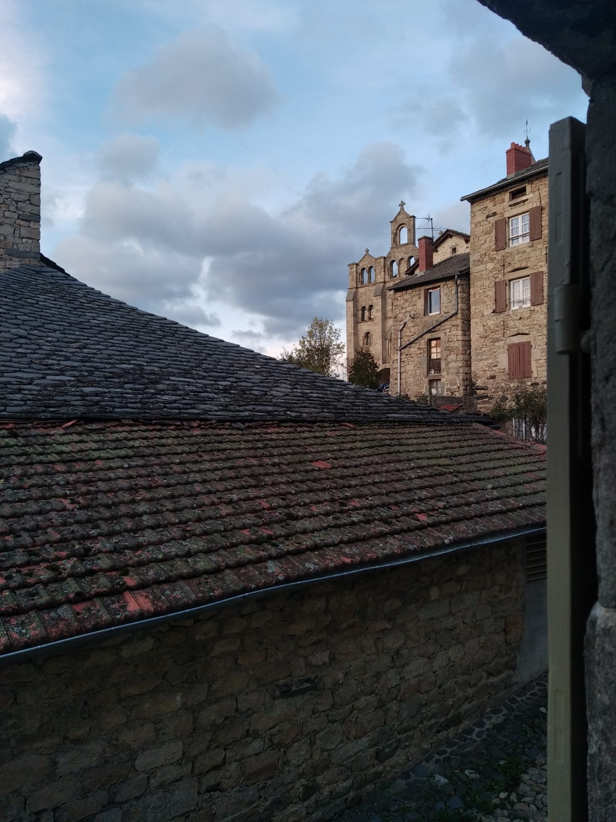 A view is captured from a window, revealing an old stone building with architectural details and a neighboring roof with textured slate tiles. Soft clouds drift across a muted sky, framing the historic surroundings.