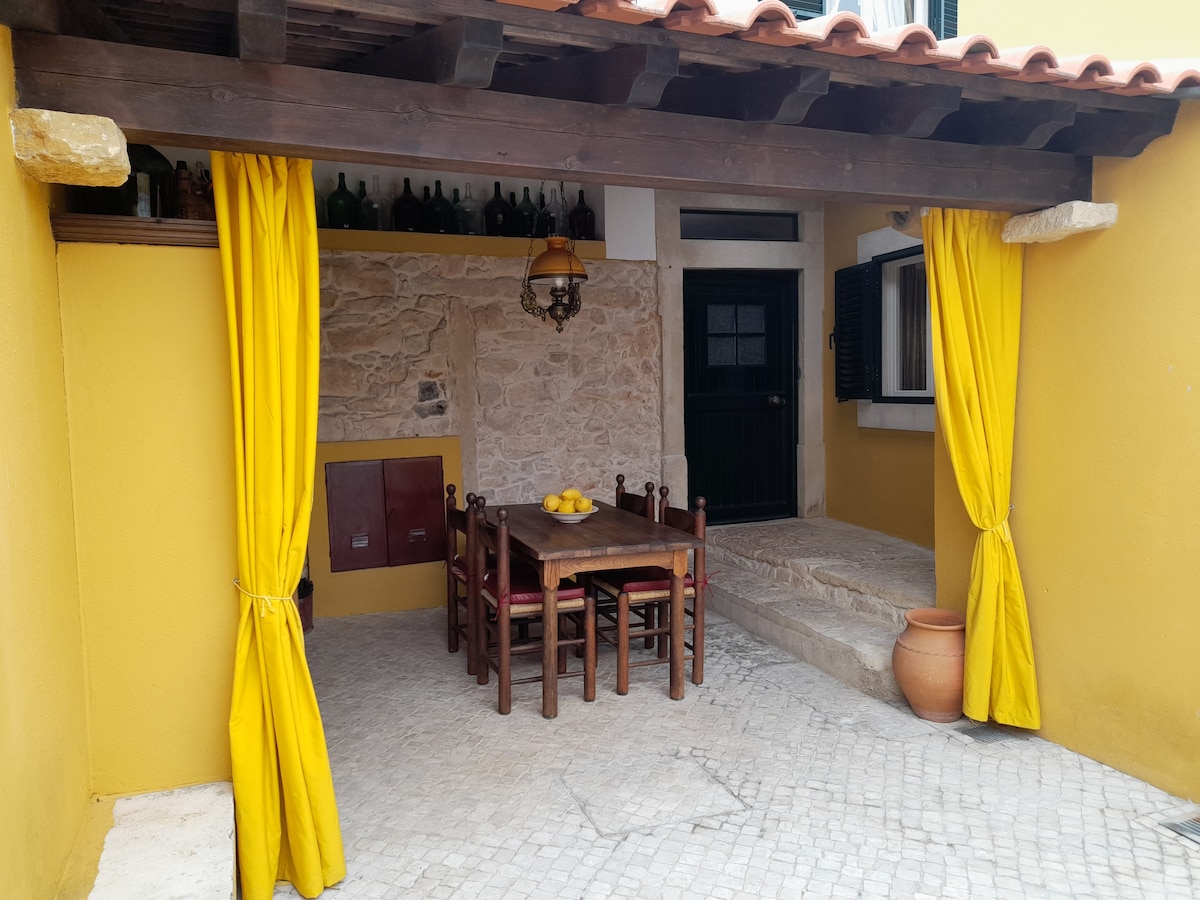 An exterior dining area is shown, featuring a wooden table with six chairs. Yellow curtains frame the space, which is highlighted by a rustic stone wall and a lantern fixture. A terracotta pot with flowers sits nearby, enhancing the inviting outdoor ambiance.