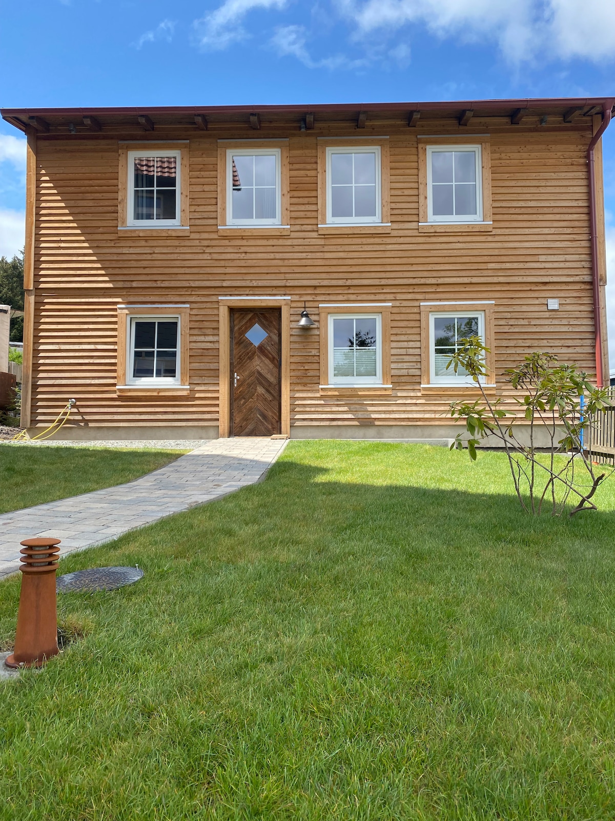 A two-story wooden house features a facade of natural wooden planks and three large windows on the front. A simple pathway leads to the entrance, which is accentuated by a wooden door with a diamond-shaped window. Lush green grass surrounds the building, complemented by a small shrub.