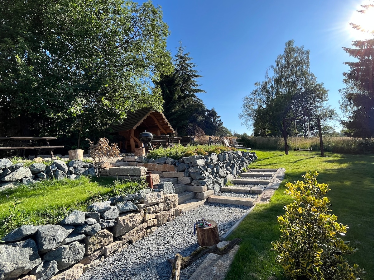 A landscaped outdoor area features natural stone steps leading through a garden with lush green grass and decorative rocks. A wooden structure is visible in the background amidst trees, complemented by seating areas and gentle sunlight filtering through. The space invites relaxation in a serene environment.