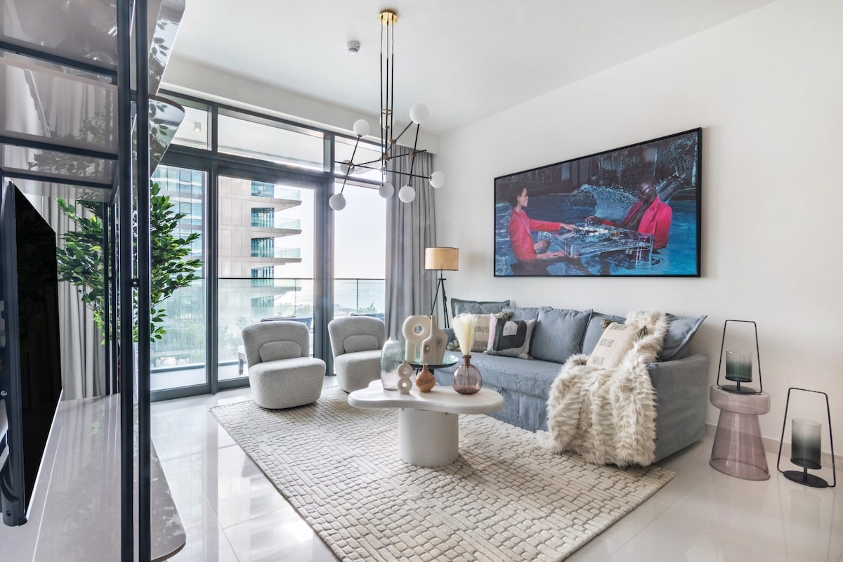 A spacious living area is highlighted by floor-to-ceiling windows, allowing natural light to fill the space. A comfortable sofa is adorned with soft throw pillows, complemented by accent chairs. A stylish coffee table sits atop a textured rug, while decorative items add a touch of elegance.