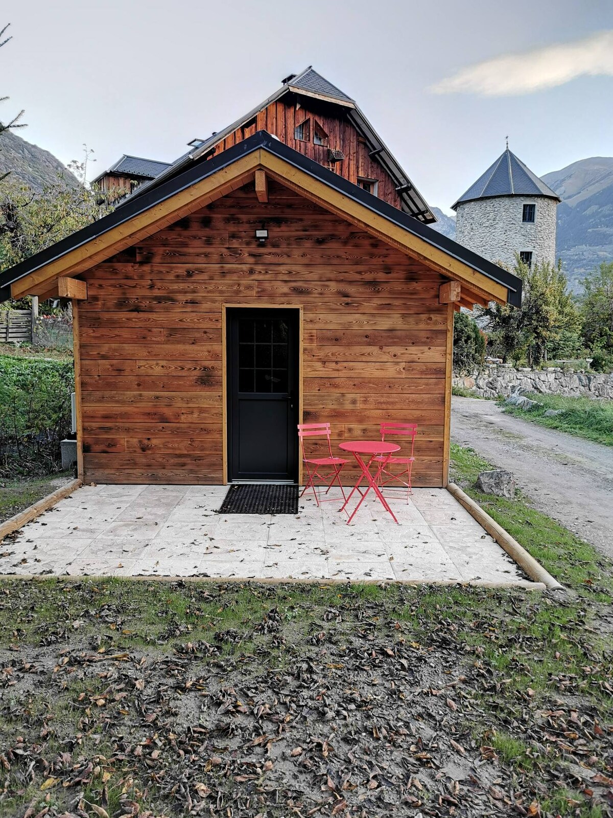 A wooden cabin features a welcoming entrance with a double door. A small, round table with pink chairs is set on the tiled patio, surrounded by a natural landscape. In the background, a stone structure and mountains are visible, completing the serene outdoor setting.