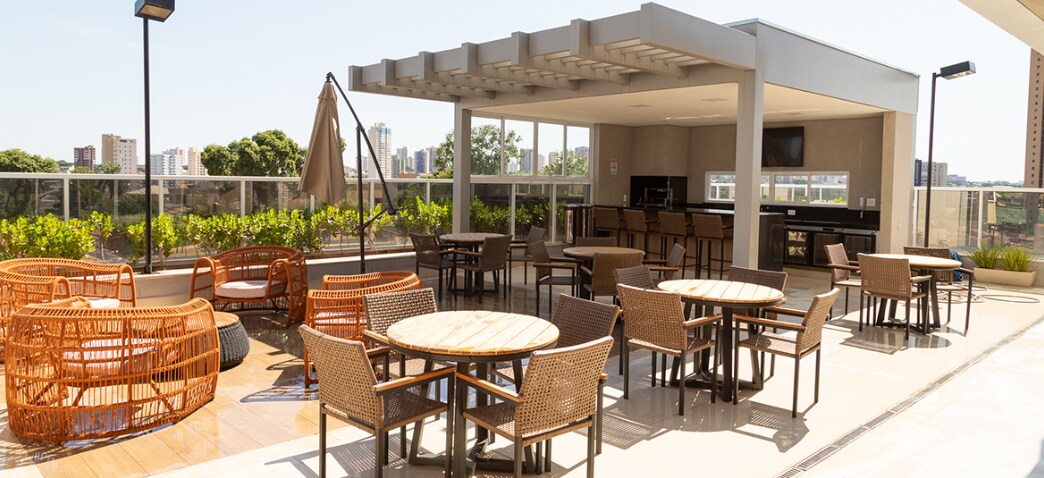 An outdoor terrace offers a variety of seating options, including circular wicker chairs and wooden tables. The area is shaded by a pergola, and a patio umbrella stands nearby. Lush greenery surrounds the space, providing a pleasant view of the city skyline.