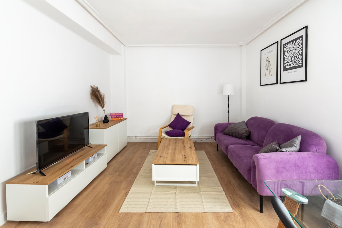 A spacious living area features a modern purple sofa and a light wood coffee table, complemented by a flat-screen TV on a sleek, white media unit. Minimalist wall art adorns the walls, creating a calm and inviting environment.