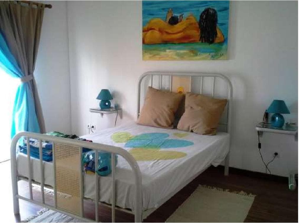A bright bedroom features a spacious bed with a colorful bedding set and two pillows. Bedside tables with teal lamps are positioned on each side. A vibrant painting hangs on the wall above the bed, and natural light enters through the window, complemented by flowing curtains.