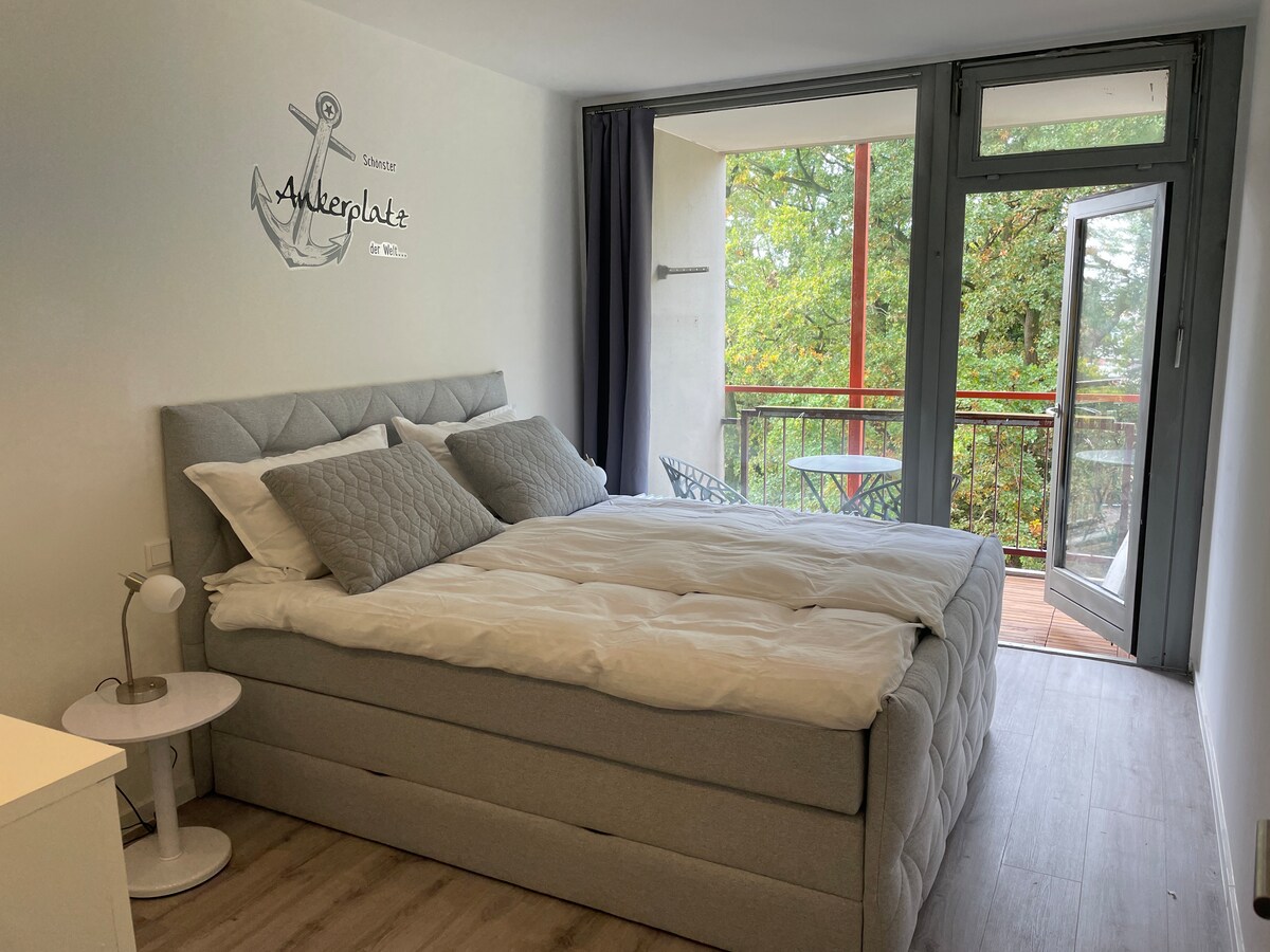 A spacious bedroom features a soft gray bed adorned with plush pillows and a light comforter. A small bedside table holds a lamp. Floor-to-ceiling windows provide access to a balcony with a small table and chairs, offering views of greenery.