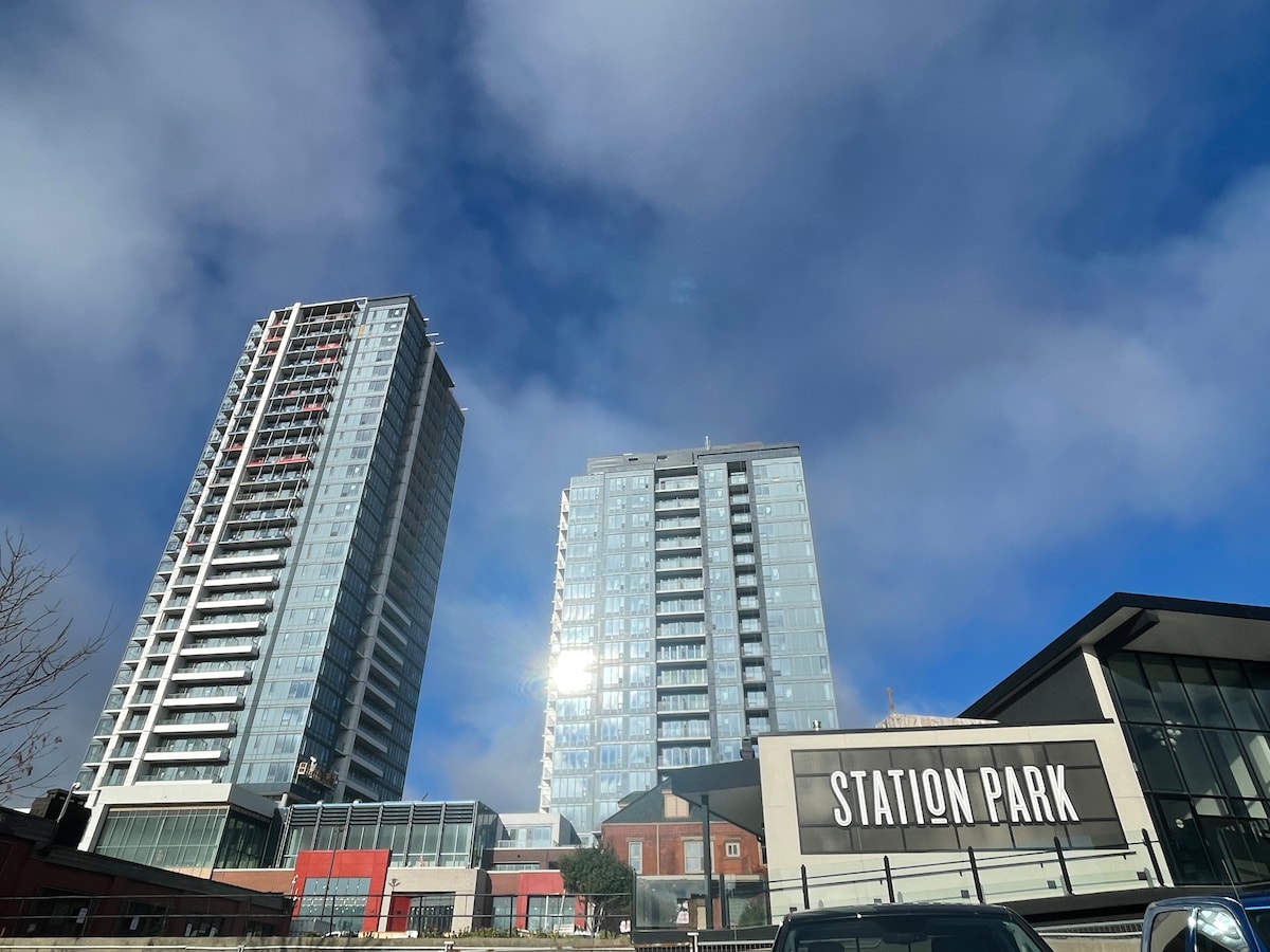 Two modern high-rise buildings are prominently featured against a blue sky with soft clouds. The buildings, part of Station Park, showcase expansive glass facades, reflecting the sunlight. A nearby sign displays the name 'STATION PARK,' indicating the development's vibrant urban setting.