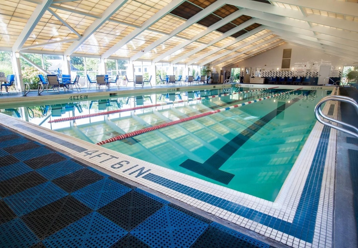 An indoor swimming pool is presented with a clear blue water surface, bordered by a safety lane line. Surrounding lounge chairs are arranged along the edges, and large windows above allow natural light to illuminate the space.