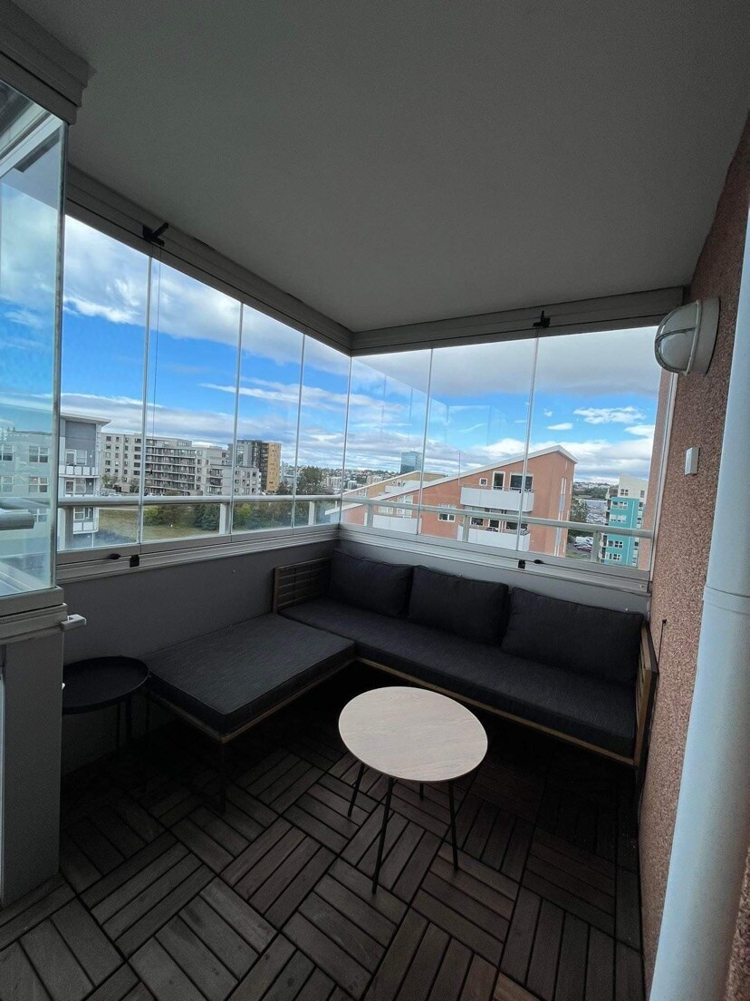 A serene balcony space is presented, featuring a dark lounge area with cushions and a small circular table. Large glass panels allow for unobstructed views of the surroundings, while wooden flooring adds warmth to the area.