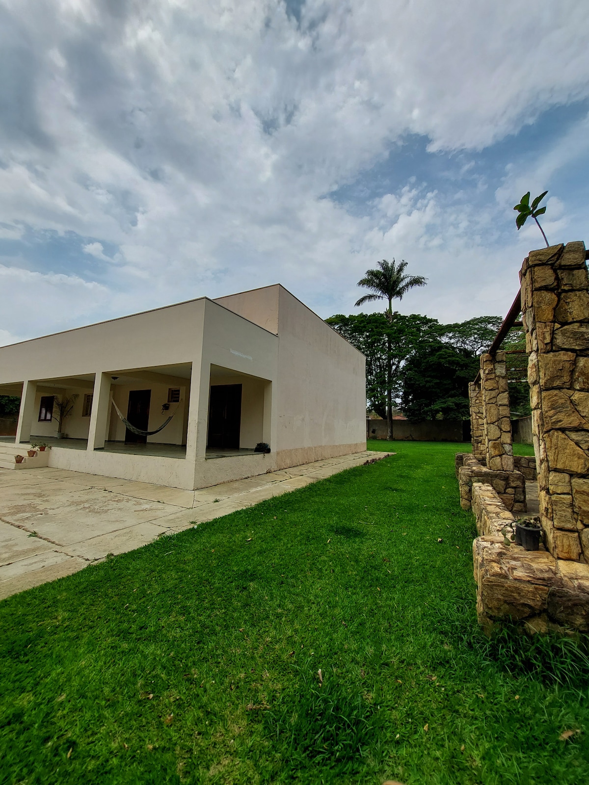 A modern one-story house is shown, featuring a minimalist design with a clean white exterior. The surrounding lawn is lush and green, accompanied by trees in the background. A stone structure is visible on the right, adding a natural element to the setting.