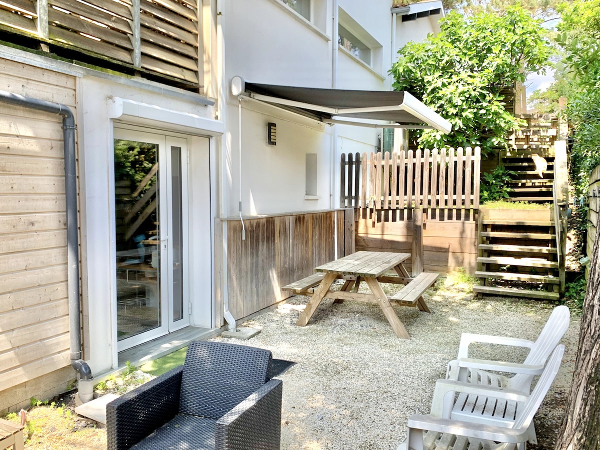 An outdoor area features a picnic table and comfortable seating arrangements. A retractable awning provides shade, while a gravel surface complements the wooden fencing and lush greenery. Steps lead to an elevated space, enhancing the private garden setting.