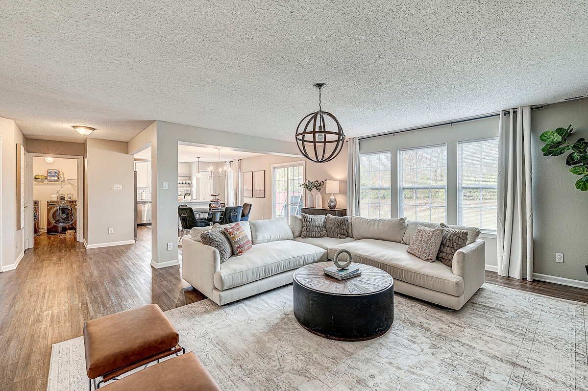 A spacious living area is showcased, featuring a light-colored sectional sofa and a round coffee table. Large windows allow natural light to illuminate the room while offering views of the serene outdoor space. A cozy throw blanket and decorative pillows add subtle comfort.