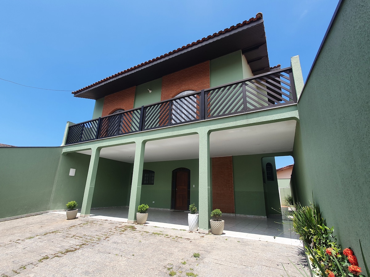 A two-story structure features a green exterior with a brick upper level. A spacious patio area is visible in the foreground, complemented by potted plants. The balcony on the upper level provides an outdoor space overlooking the entrance.