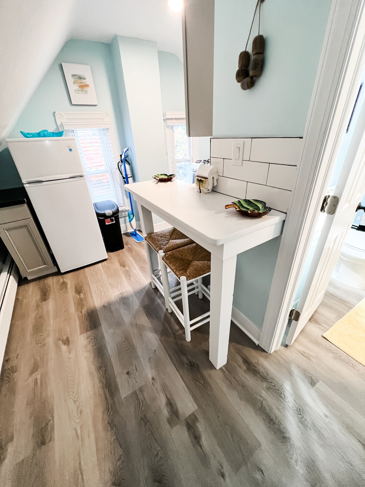 A compact kitchen area is accentuated by light blue walls and warm wooden flooring. A small white table with two woven stools is positioned near a window, allowing natural light to enter. A refrigerator stands adjacent to the countertop, creating a functional space.
