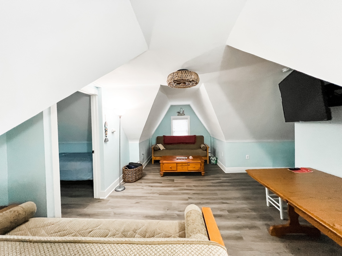 An attic living space features sloped ceilings with soft blue walls, providing a cozy ambiance. A comfortable sofa and a wooden table are arranged for relaxation or dining. A television is mounted on the wall, and a glimpse of a separate sleeping area is visible through an open door.