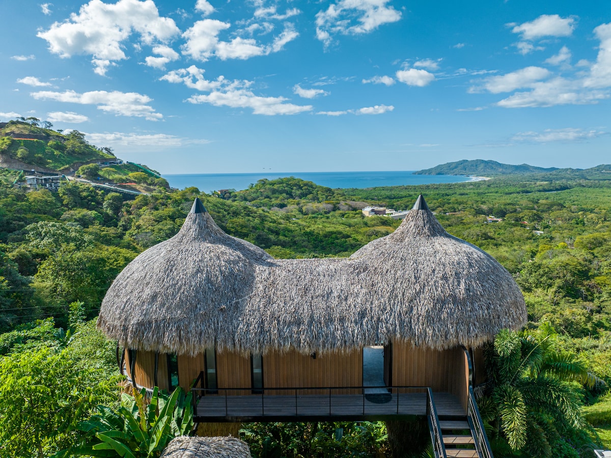 The villa is elevated, featuring a distinctive thatched roof and wooden exterior that harmonizes with its natural surroundings. Verdant hills and ocean views are visible in the background, creating a tranquil atmosphere framed by a clear blue sky.