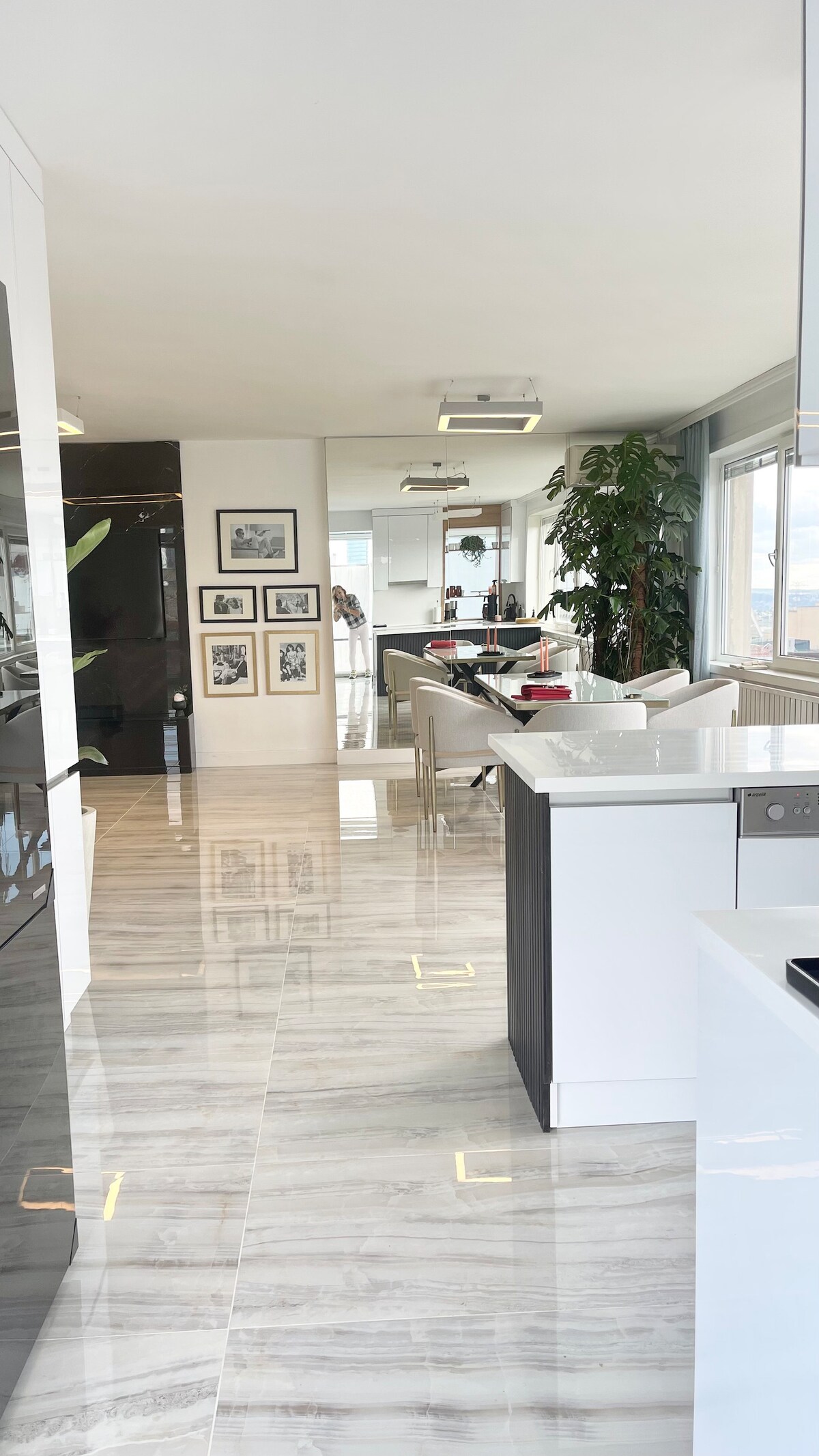 A spacious and modern kitchen is visible, featuring sleek cabinetry and a white countertop. Natural light floods the area, highlighting the glossy marble flooring. In the background, a dining space is seen with multiple chairs, while greenery adds a touch of vibrant color to the room.