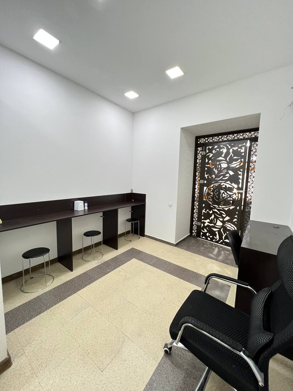 A well-lit room features a sleek entrance door with intricate designs. Two high stools are positioned at a dark wooden counter, while a black office chair is placed against the wall. Neutral-toned flooring contrasts with the dark furniture and bright overhead lights.