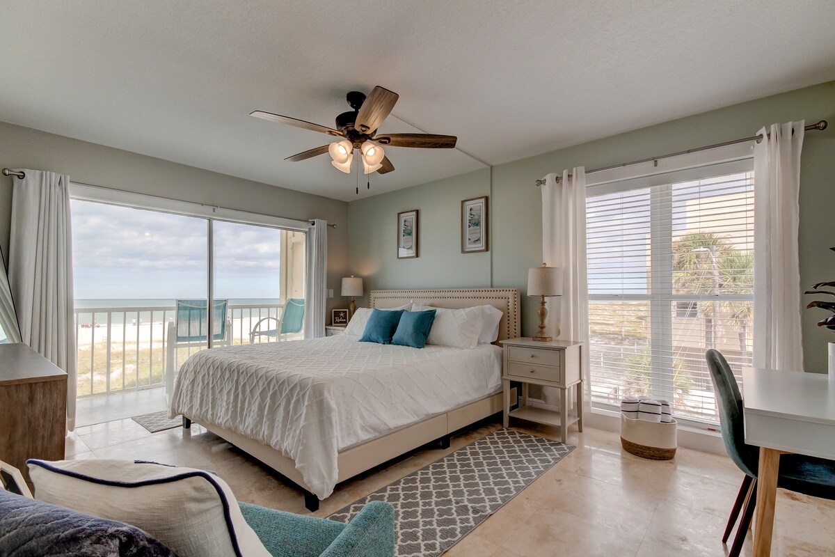A spacious bedroom features a comfortable bed with light bedding and decorative pillows. Large windows provide views of the beach, while a balcony is accessible from the room. Soft natural light brightens the area, complemented by a ceiling fan and minimalist decor.