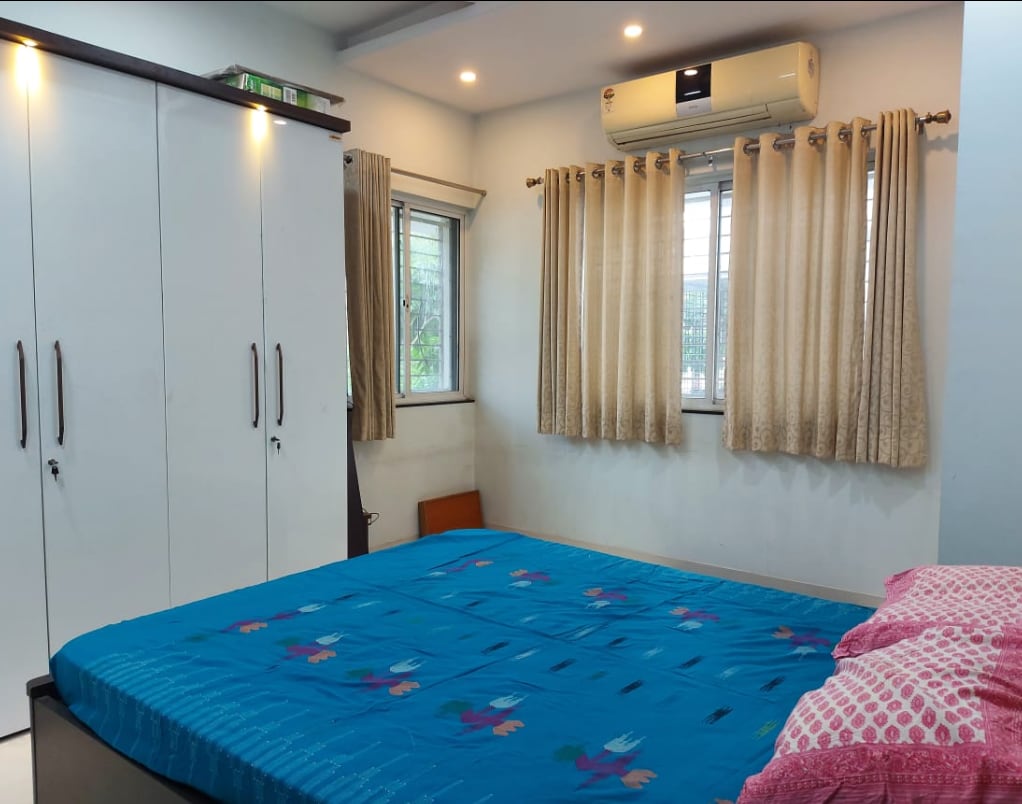 A bedroom is furnished with a spacious bed covered by a blue floral bedspread. Light-colored curtains frame the windows, allowing natural light to enter. A built-in wardrobe is visible on one side, and an air conditioning unit is mounted above the bed.