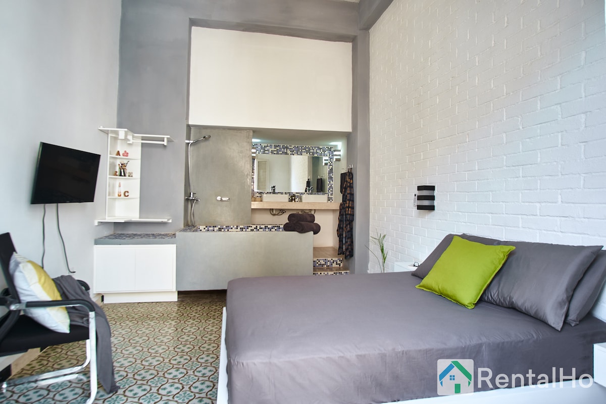 A spacious room features a minimalist design with a large bed and a decorative green pillow. A wall-mounted TV is installed opposite the bed, while a modern sink area is visible in the background. The patterned floor adds character to the space.