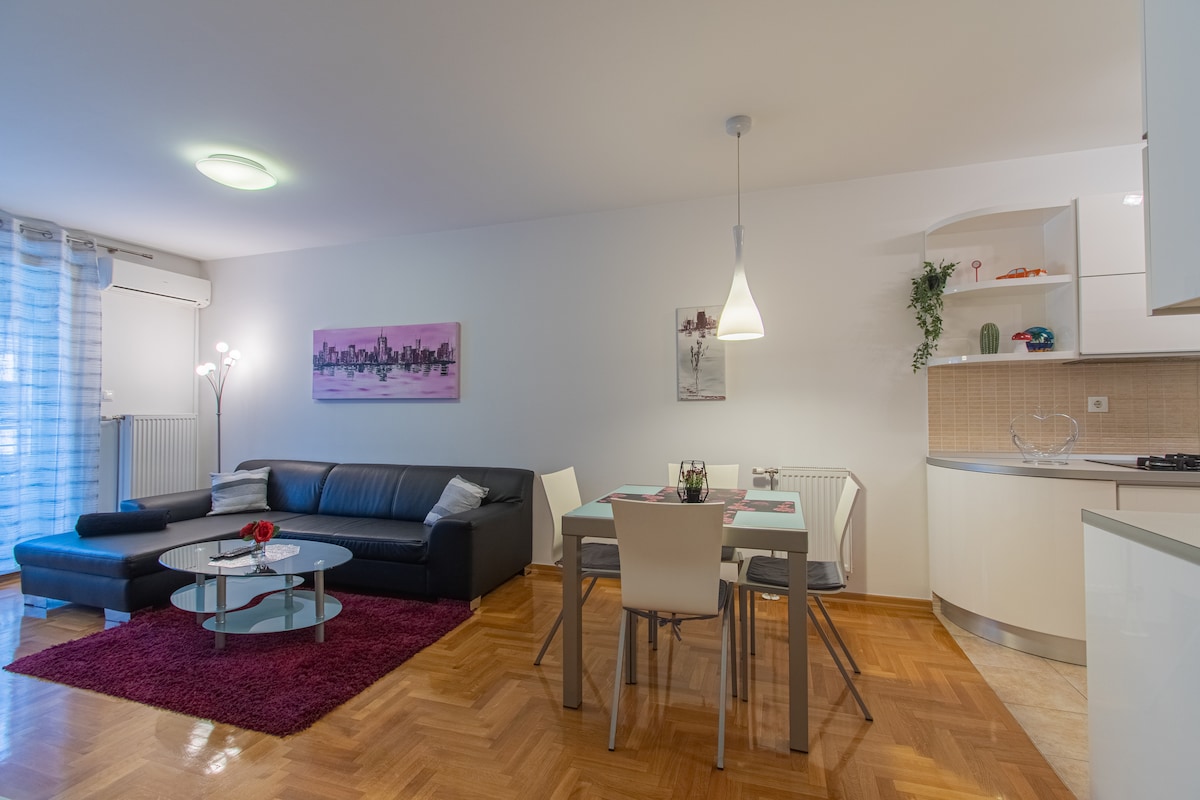 A modern living area features a dark sectional sofa and a glass coffee table resting on a plush area rug. A dining table with four chairs sits beside the kitchenette. Soft lighting and artwork on the walls create a pleasant ambiance.