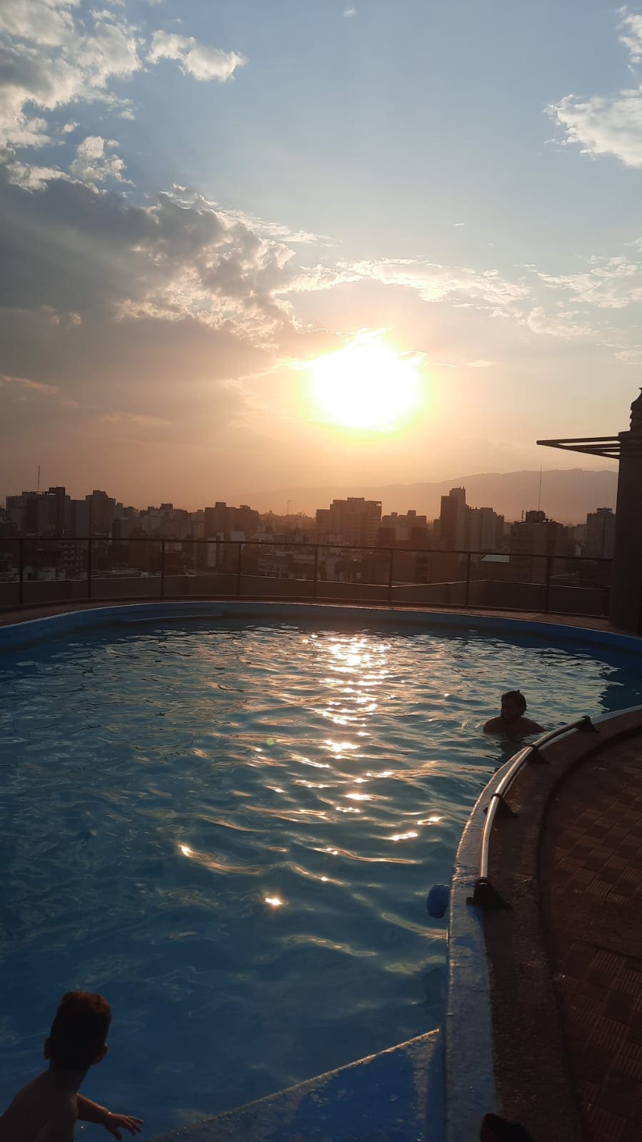 A rooftop pool is featured, reflecting the golden tones of a sunset. The city skyline is visible in the background, with various buildings outlined against the colorful sky. Calm water creates a serene atmosphere, inviting relaxation.
