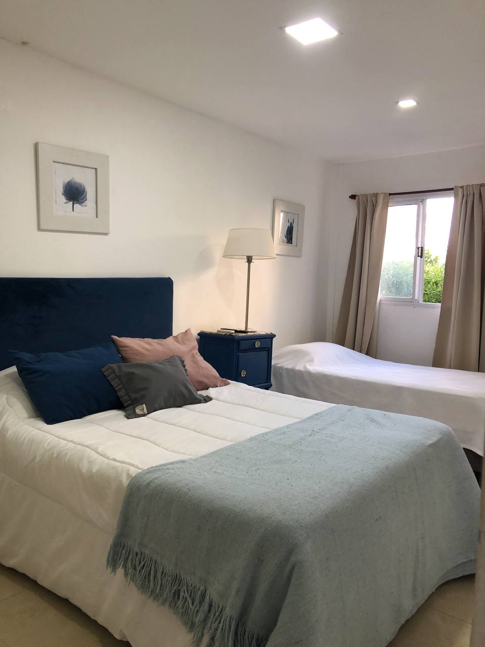 A bright sleeping area features two beds, one with a soft blue headboard and decorative pillows. A light blanket is draped over the larger bed. Natural light enters through a window covered by sheer curtains, illuminating the space, which is complemented by framed wall art.