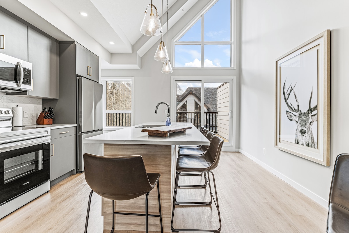 A modern kitchen area features stainless steel appliances and an island with seating for four. Large triangular windows allow natural light to fill the space, highlighting the sleek cabinetry. A framed artwork depicting a deer is visible on the wall, adding a decorative touch.