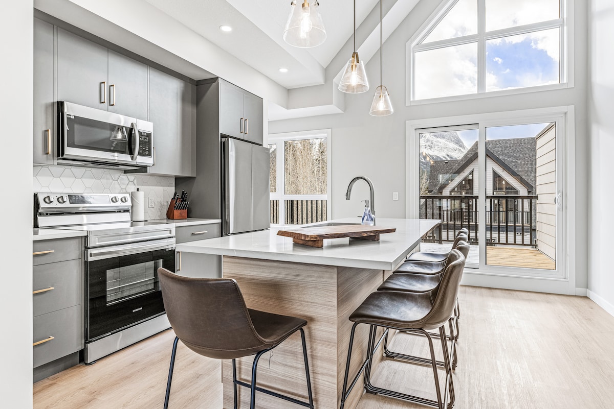A modern kitchen features stainless steel appliances, including a microwave and oven. An island with seating for five is centrally positioned, illuminated by hanging pendant lights. Large windows allow natural light to fill the space, with doors leading to a private outdoor area.