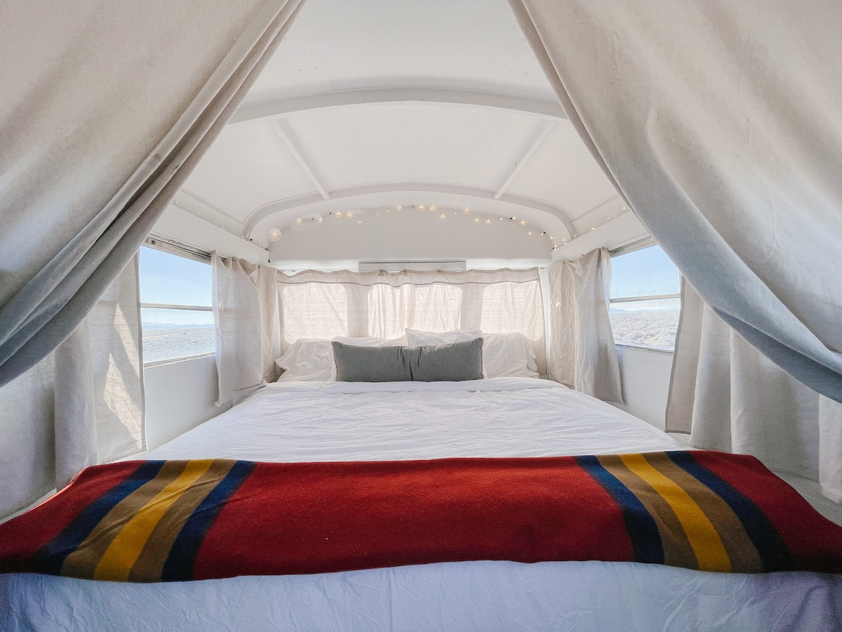 A spacious king-sized bed is highlighted with premium linens and decorative pillows, set beneath soft curtains. String lights adorn the ceiling, creating a cozy ambiance. Natural light floods the interior, enhancing the minimalistic design and inviting a serene retreat.