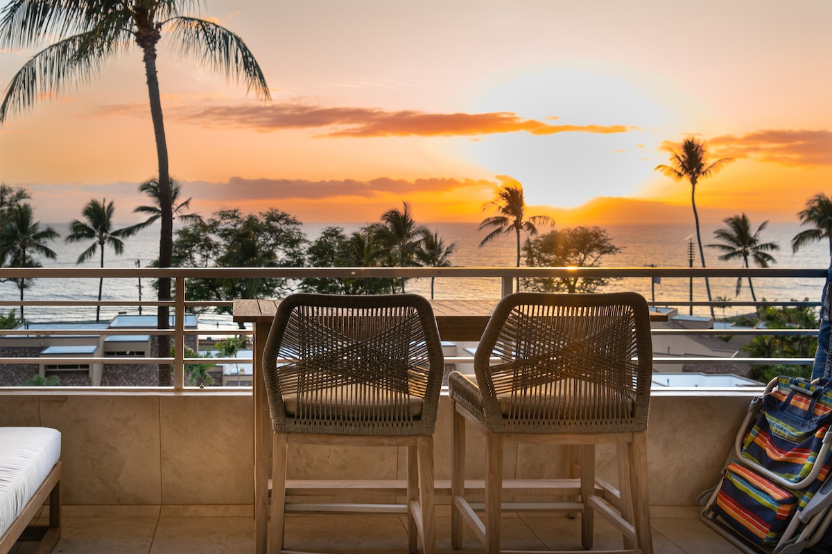 A serene lanai features two counter-height chairs facing the ocean, providing unobstructed sunset views. Lush palm trees frame the backdrop, while a colorful beach bag rests on the side. The tranquil atmosphere invites relaxation amidst the natural beauty of Maui.