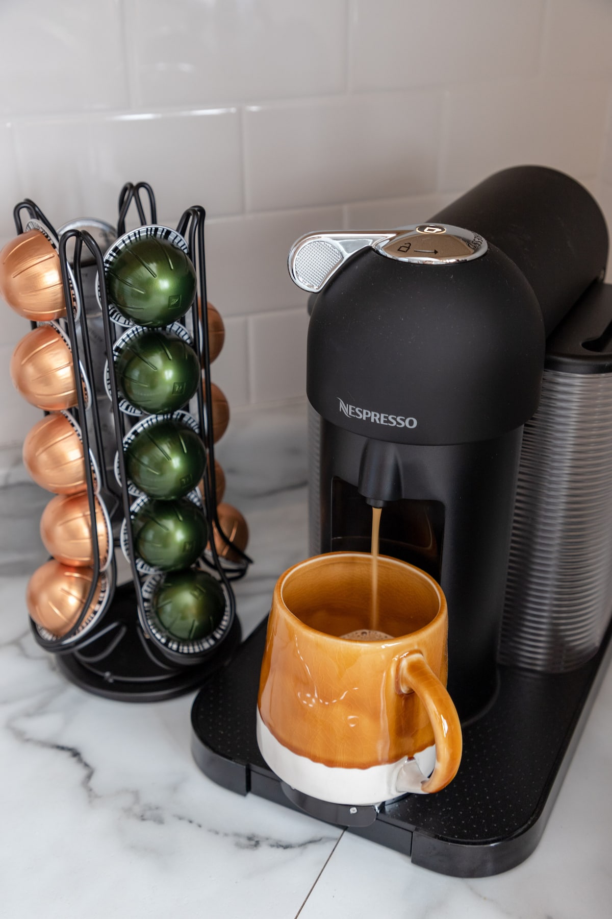 An espresso machine is situated on a marble countertop, with a warm-toned mug placed beneath its spout. A vertically arranged rack stands next to it, displaying colorful coffee capsules in shades of green and copper.
