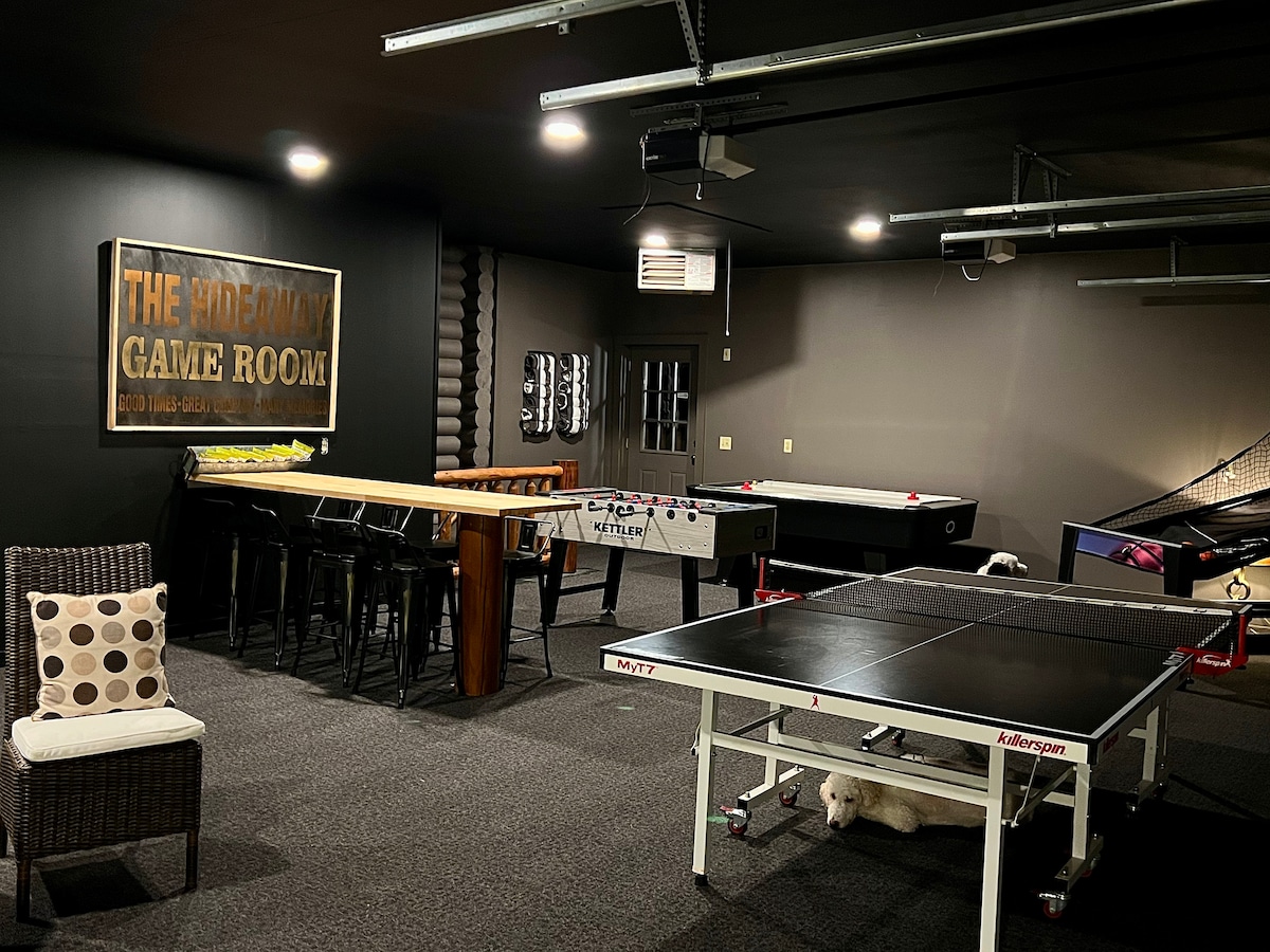 The game room presents an engaging environment with various activity options. A foosball table, ping pong table, and air hockey table are visible alongside a seating area that includes a chair with decorative pillows. A bar-height table is stationed nearby, enhancing the space for social interactions.