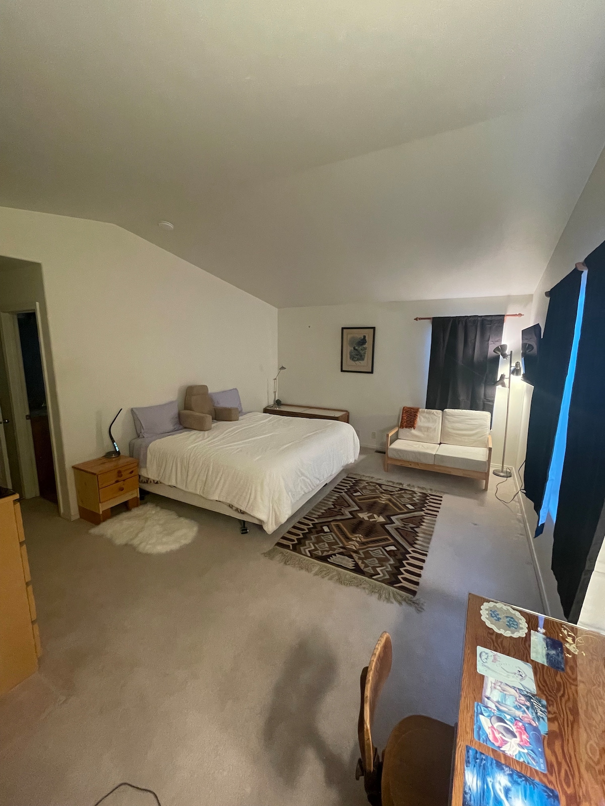 A spacious bedroom features a large bed with a light-colored duvet and two accent pillows. A cozy seating area includes a white lounge chair beside a wooden desk. The room is adorned with a textured area rug, and windows with dark curtains allow for light control.