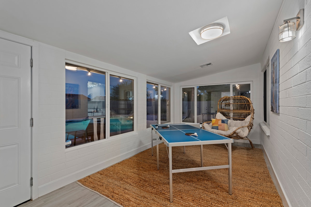 An air-cooled game room is featured with two ping pong tables positioned near large windows, providing views of the heated pool outside. A comfortable hanging chair adds seating. The space is completed with a neutral-colored area rug, enhancing the bright and open atmosphere.