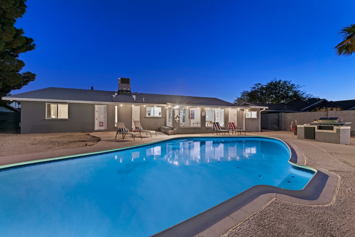 The private backyard features a sparkling heated pool, surrounded by lounge chairs and accessible outdoor lighting. A custom BBQ island is visible, adding to the inviting space for gatherings. The home's exterior, with a mid-century modern design, is illuminated, creating a welcoming atmosphere.